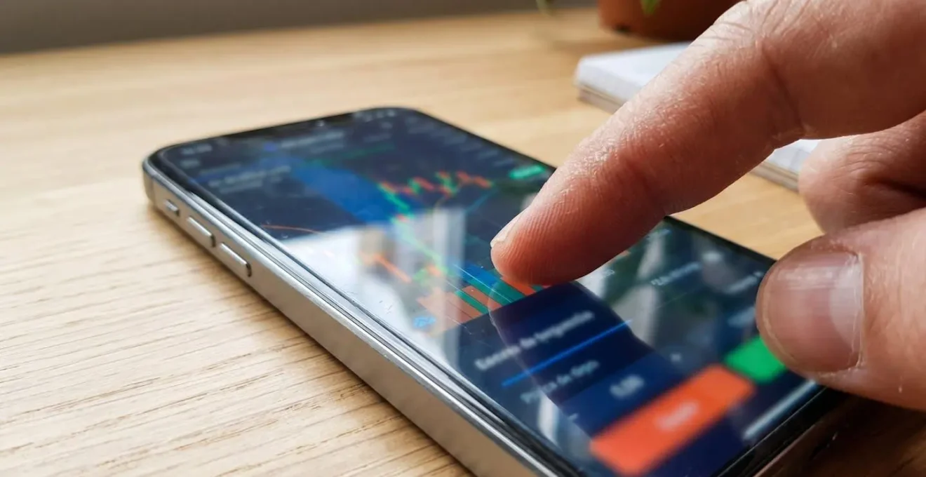 Primer plano de un dedo tocando la pantalla de un smartphone que muestra un gráfico borroso de evolución de precios, sobre superficie de madera moderna
